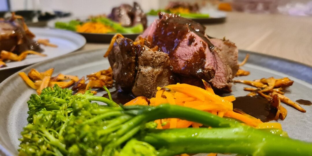 En tallerken med mørt kjøtt, servert med stekt sopp, friske grønnsaker som brokkolini og gulrotstrimler, og toppet med en rik brun saus. Retten er pent presentert på en grå tallerken