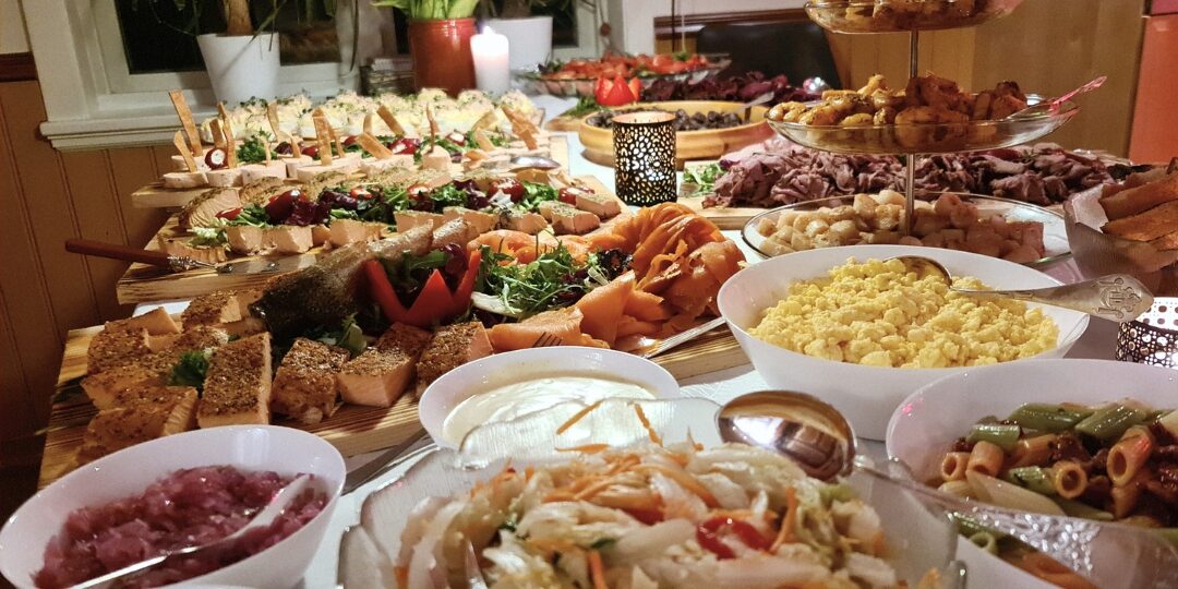 Et bugnende buffetbord fylt med et utvalg av retter, inkludert røkelaks, salater, pasta, brød og ulike kjøttretter, dekorert med levende lys og presentert i et koselig innemiljø