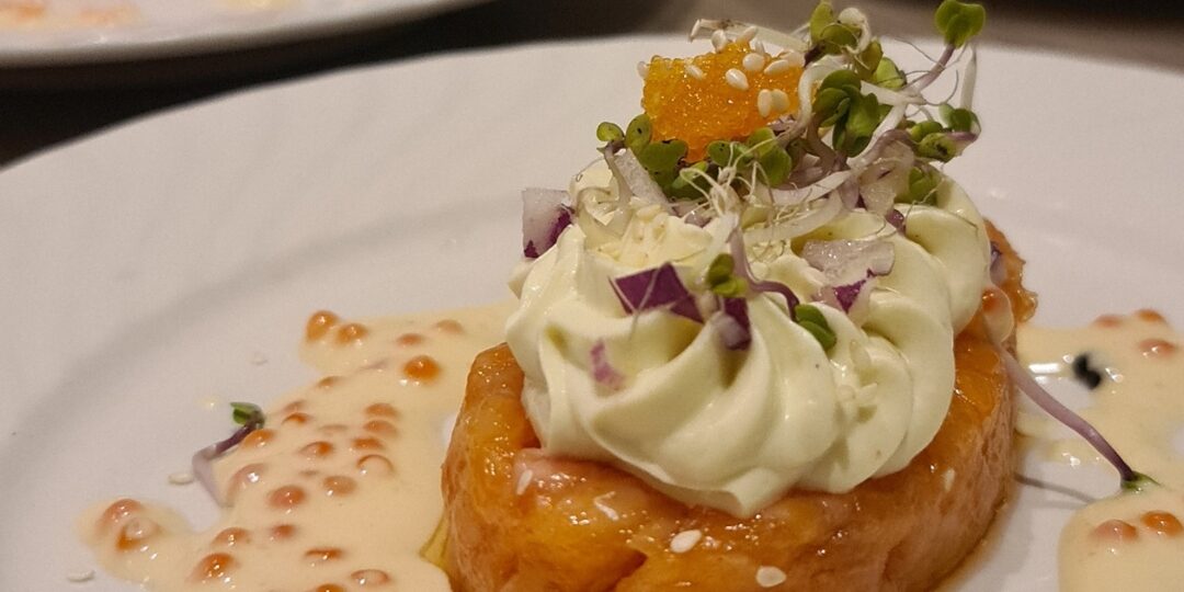 En delikat forrett bestående av laksetartar toppet med kremet dressing, små spirer, rogn og sesamfrø, servert med en lett saus på en hvit tallerken. Flere porsjoner av retten vises i bakgrunnen, klar til servering