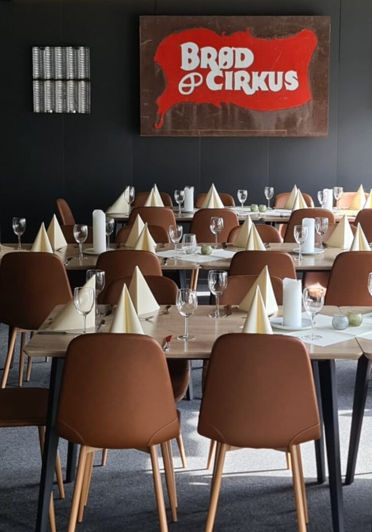 Et elegant selskapslokale med enkle, stilrene bordoppsett. Bordene er dekket med hvite tallerkener, glass og sammenbrettede servietter, mens stolene har en brun lærfinish. I bakgrunnen henger et skilt med teksten ‘Brød & Circus’, og lokalet har en rustikk atmosfære med mursteinsdetaljer og moderne dekorasjoner